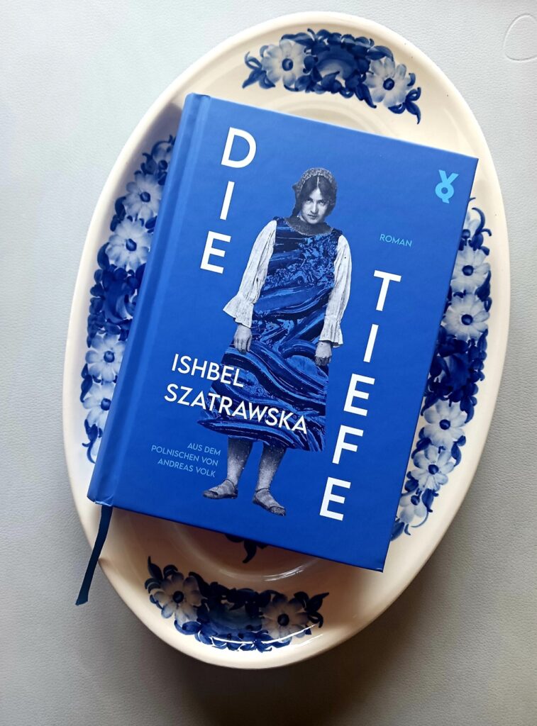 Foto des Buches »Die Tiefe« von Ishbel Szatrawska, auf dessen blauem Cover eine junge Frau mit traditioneller blauer Kleidung und Haube steht. Sie lässt die Arme hängen und schaut von unten in die Kamera. Das Buch liegt auf einer mit blauen Blumen bemalten weißen Servierplatte. Lektorat