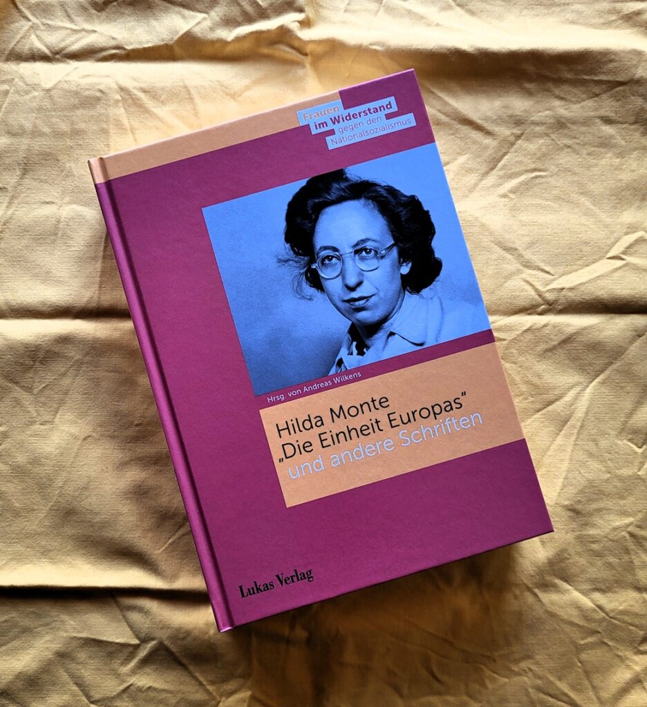 Foto des Buches Hilda Monte, »Die Einheit Europas« und andere Schriften, herausgegeben von Andreas Wilkens. Das Buch, dessen Cover in Orange und Rot gehalten ist, zeigt ein Schwarz-Weiß-Porträt der erwachsenen Hilda Monte; sie trägt Brille, hat die Haare locker zusammengesteckt und schaut ernst in die Kamera. Das Bild liegt auf gelbem Stoff. Lektorat