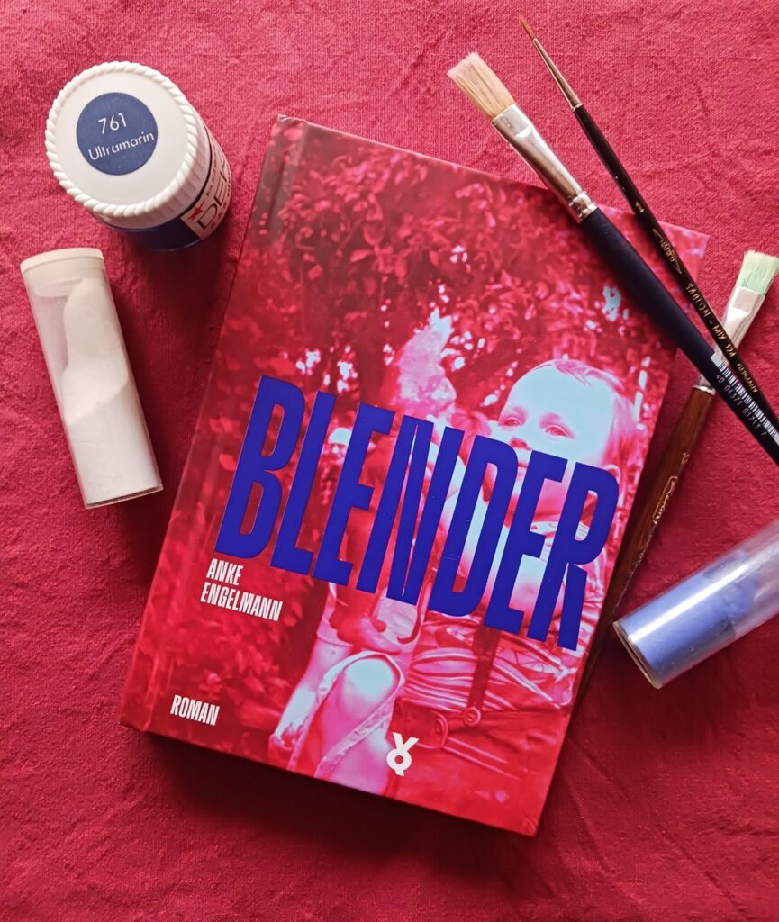 Foto des Buches. Das Cover ist rot mit blauer Schrift, es zeigt das farbverfremdete Foto eines kleinen Jungen, der einen Gartenzwerg in die Luft hält und anstrahlt. Das Buch liegt auf rotem Stoff, um es herum sind zwei Behälter mit Farbpigmenten (weiß und blau), ein Farbtopf (Farbbezeichnung ultramarin) sowie drei Pinsel drapiert.