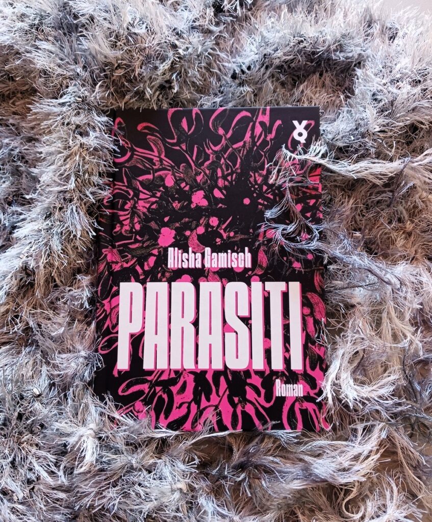 Das Buch »Parasiti« von Alisha Gamisch, auf dessen Cover eine pink-schwarzes Geschling, eventuell eine Pflanze oder ein Wesen mit so etwas wie Tentakeln, zu sehen ist, liegt in ein grau-weißen sehr flauschigen Schal und wird von allen Seiten von dessen Fransen »bedroht«.