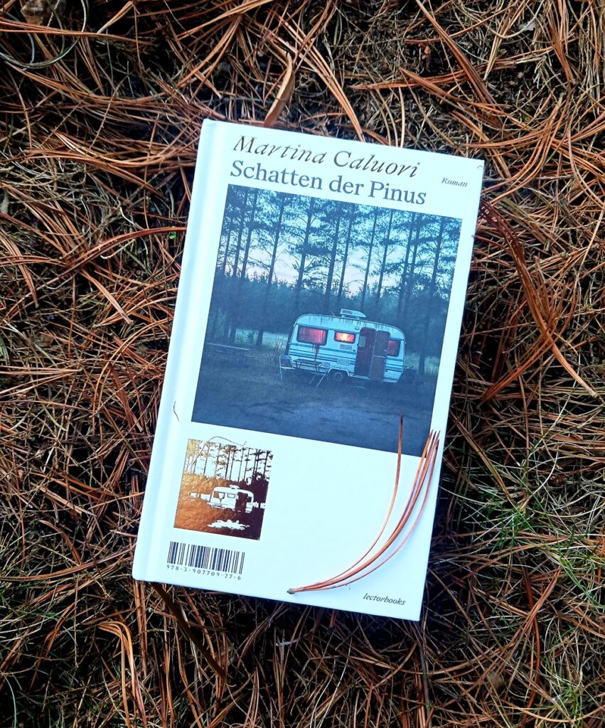 Das Buch »Schatten der Pinus« von Martina Caluori, auf dessen weißem Cover das Foto eines weißen runden Campinganhängers zu sehen ist. Er steht in der Abenddämmerung vor einer Reihe lichter Kiefern, hinter seinen Fenstern brennt Licht, vor ihm steht ein Campingtisch mit zwei Stühlen. Das Buch liegt auf braunen Kiefernnadeln, eine hat sich auf das Buch »verirrt«. Lektorat