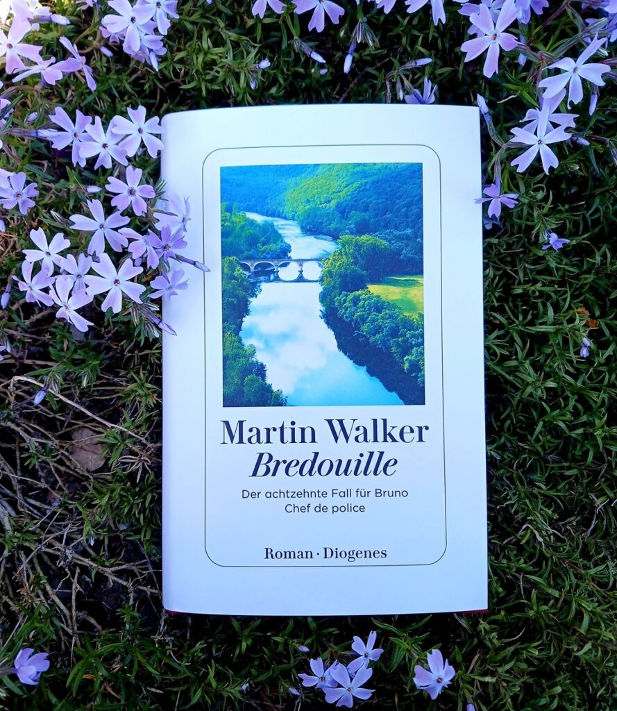 Foto des Buches »Bredouille« von Martin Walker, das in typischen Diogenes-Gestaltung ein Foto eines Flusses zeigt, der durch eine Grüne Landschaft fließt. Im Wasser spiegeln sich der Himmel und weiße Wölkchen, eine Brücke mit mehreren Bögen überspannt ihn. Das Buch liegt auf hellviolett blühendem Phlox. Die Lektorin hat das Übersetzungslektorat übernommen.