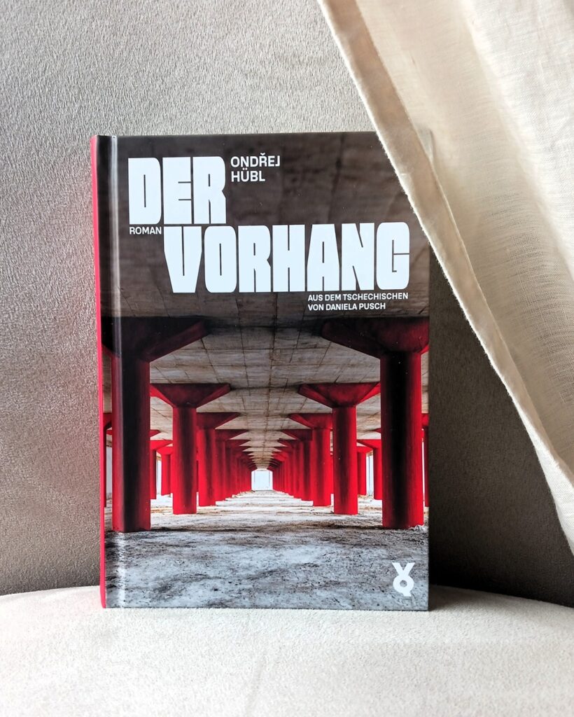Foto des Buches »Der Vorhang« von Ondřej Hübl. Auf dem Cover eine Flucht an rot gestrichenen Säulen, die auf eine weiße helle Fläche zuläuft; die Säulen stützen eine rohe Betondecke; rechts und links der Flucht sind weitere Säulen zu sehen – vielleicht ein Parkdeck oder eine Unterführung. Das Buch steht auf und vor einer hellen Stofffläche, rechts ragt ein heller Vorhang ins Bild, der die rechte obere Ecke des Buches verdeckt. Übersetzungslektorat
