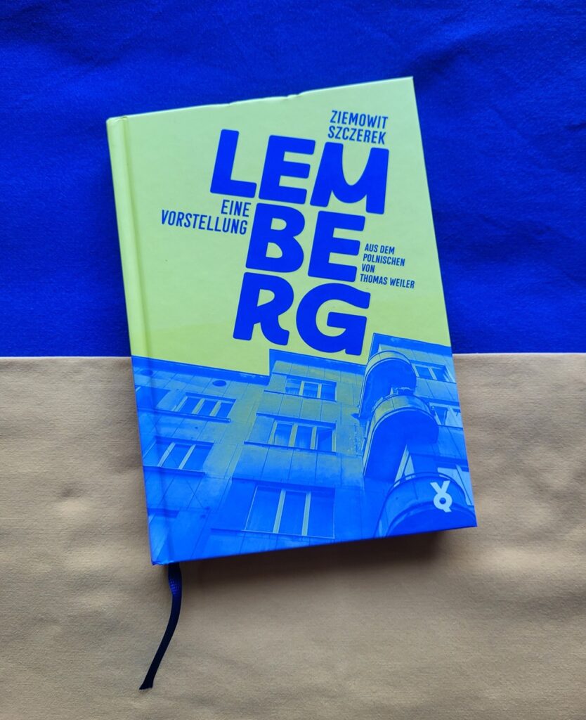 Foto des Buches »Lemberg« von Ziemowit Szczerek, dessen Cover die etwas knalligeren Farben der ukrainischen Fahne in umgekehrter Anordnung zeigt – oben gelb, unten blau, wobei das Blau drei Stockwerke einer schräg von unten aufgenommenen Hausfassade zeigt, die neben drei Vorsprüngen markante runde Balkone aufweist; der Himmel ist gelb. Das Buch liegt auf den Farben der ukrainischen Fahne.